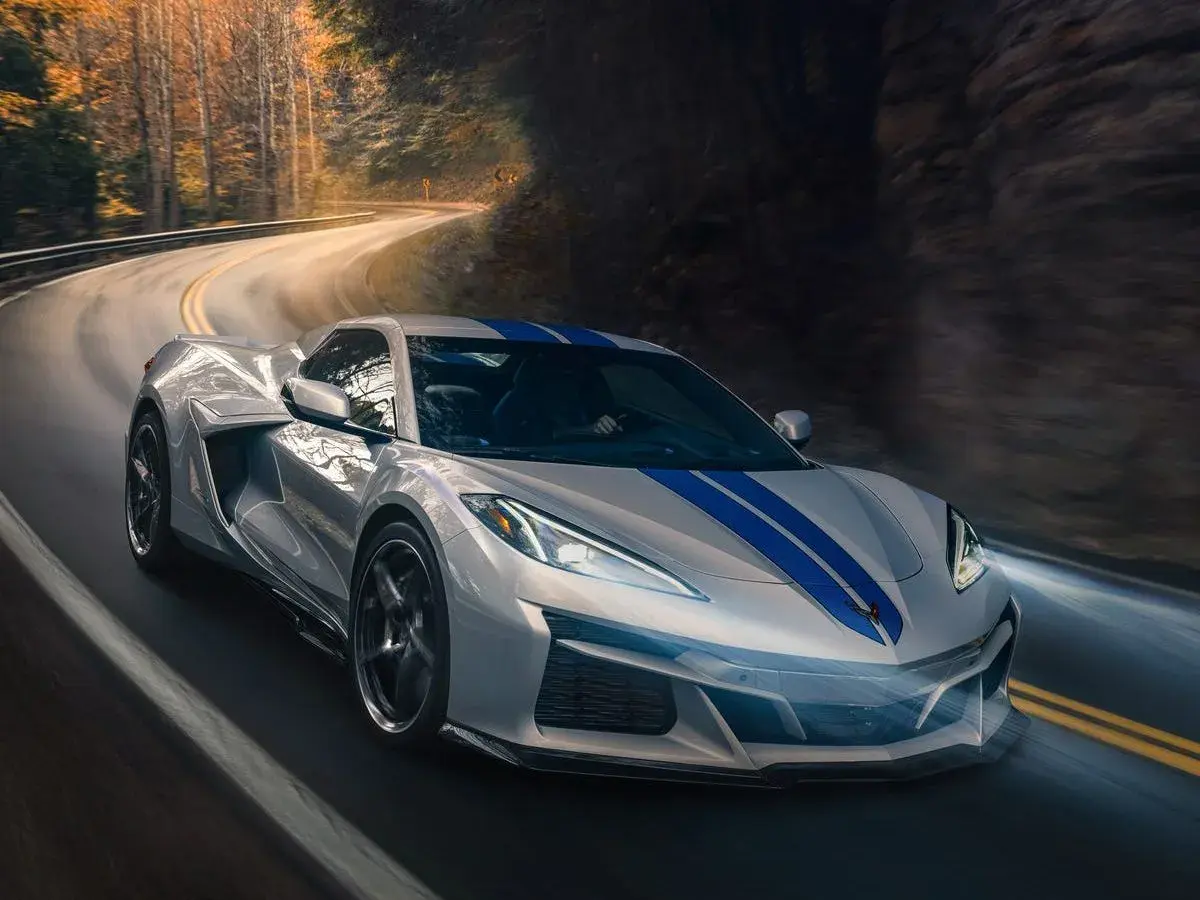 Un deportivo plateado con franjas azules recorre una carretera sinuosa entre árboles. Podría ser el mejor coche del mundo.