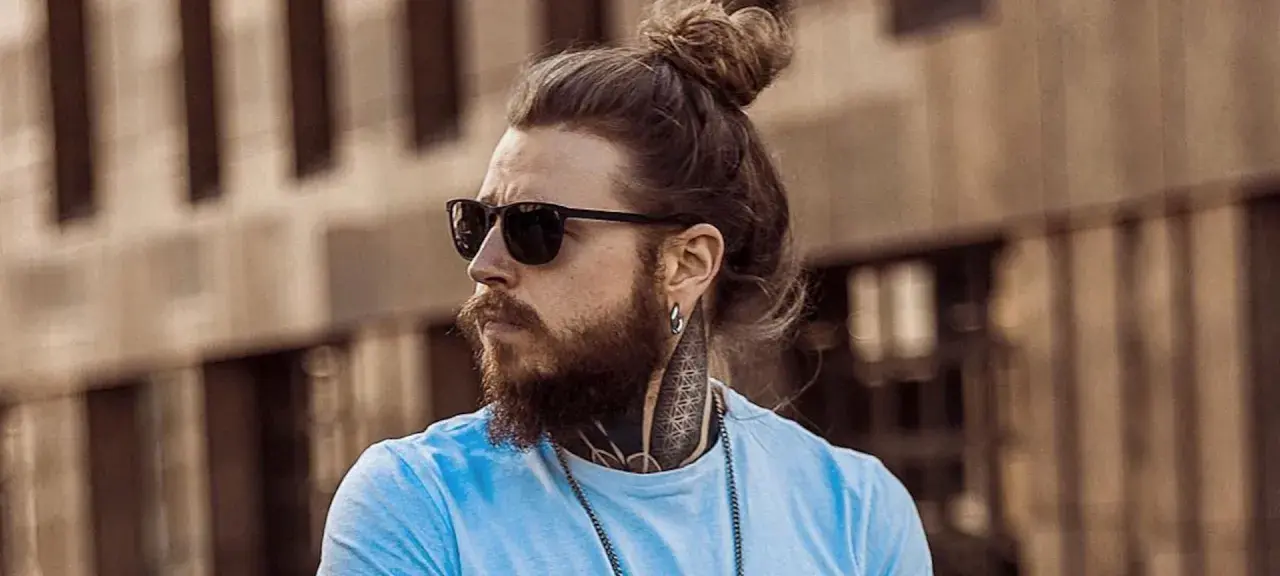 Hombre con barba y pelo recogido en moño, luciendo gafas de sol. Su estilo muestra una transicion pelo corto a largo, con tatuajes visibles en el cuello.