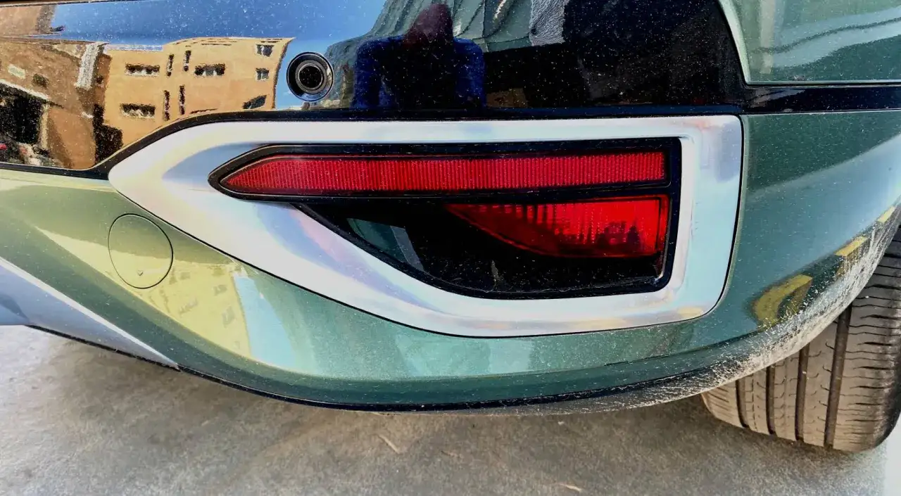 Detalle de un coche con luz trasera roja y un elemento plateado. El reflejo muestra un edificio. Este es un ejemplo de catadioptrico que es un reflector.