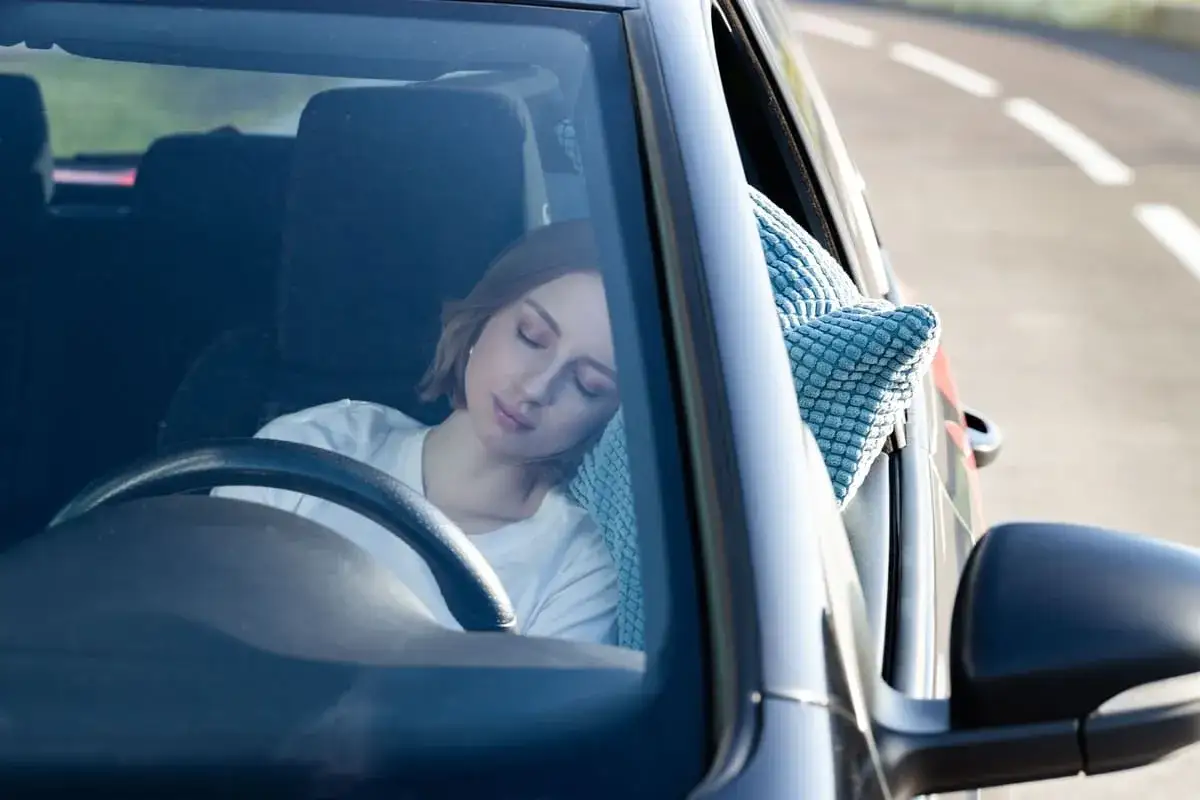 Mujer rubia duerme en el asiento del conductor, apoyada en una almohada azul. Se puede dormir en el coche en un viaje largo.