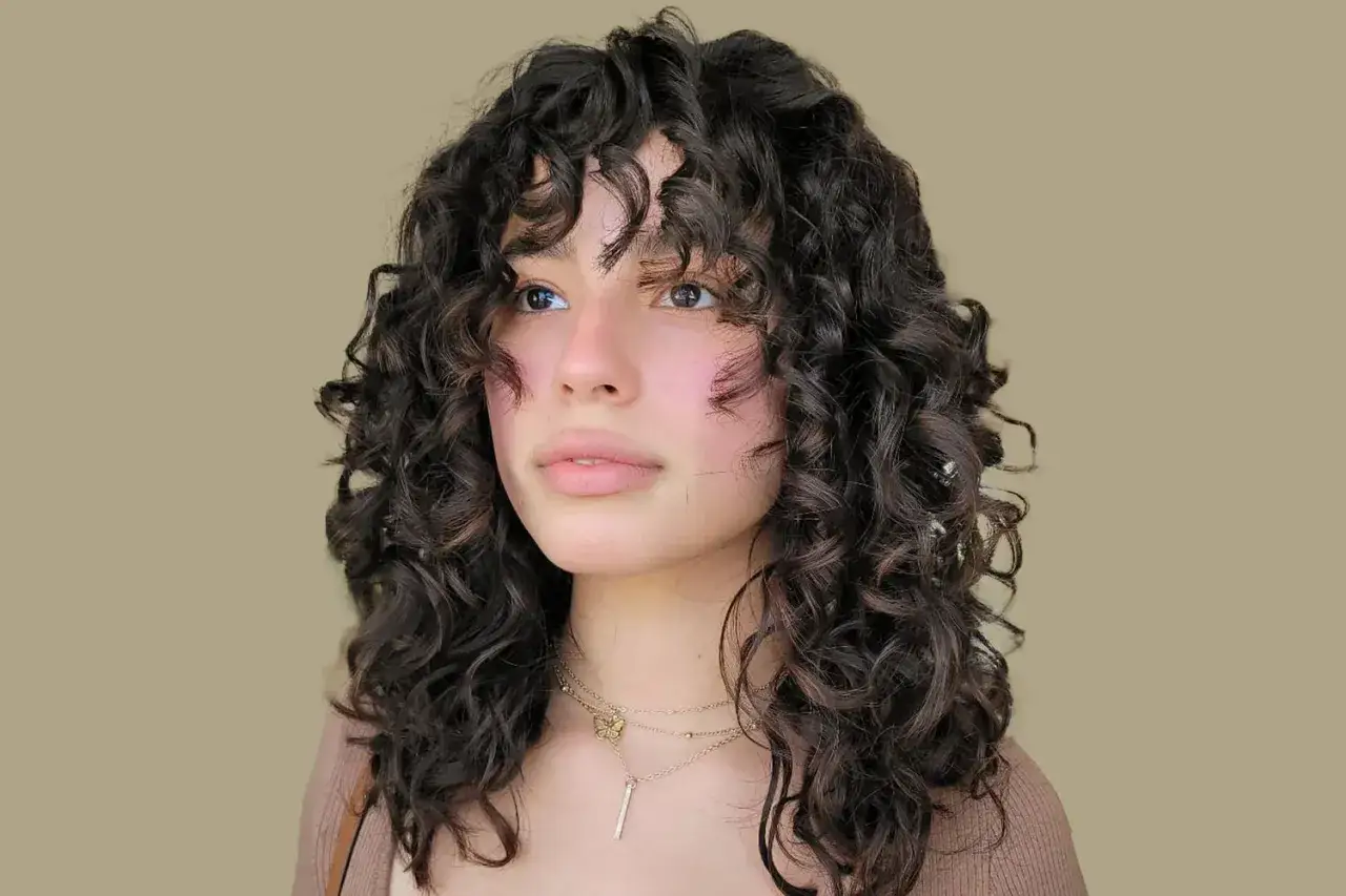 Chica con flequillo pelo rizado oscuro, mirada pensativa, luciendo collares dorados.