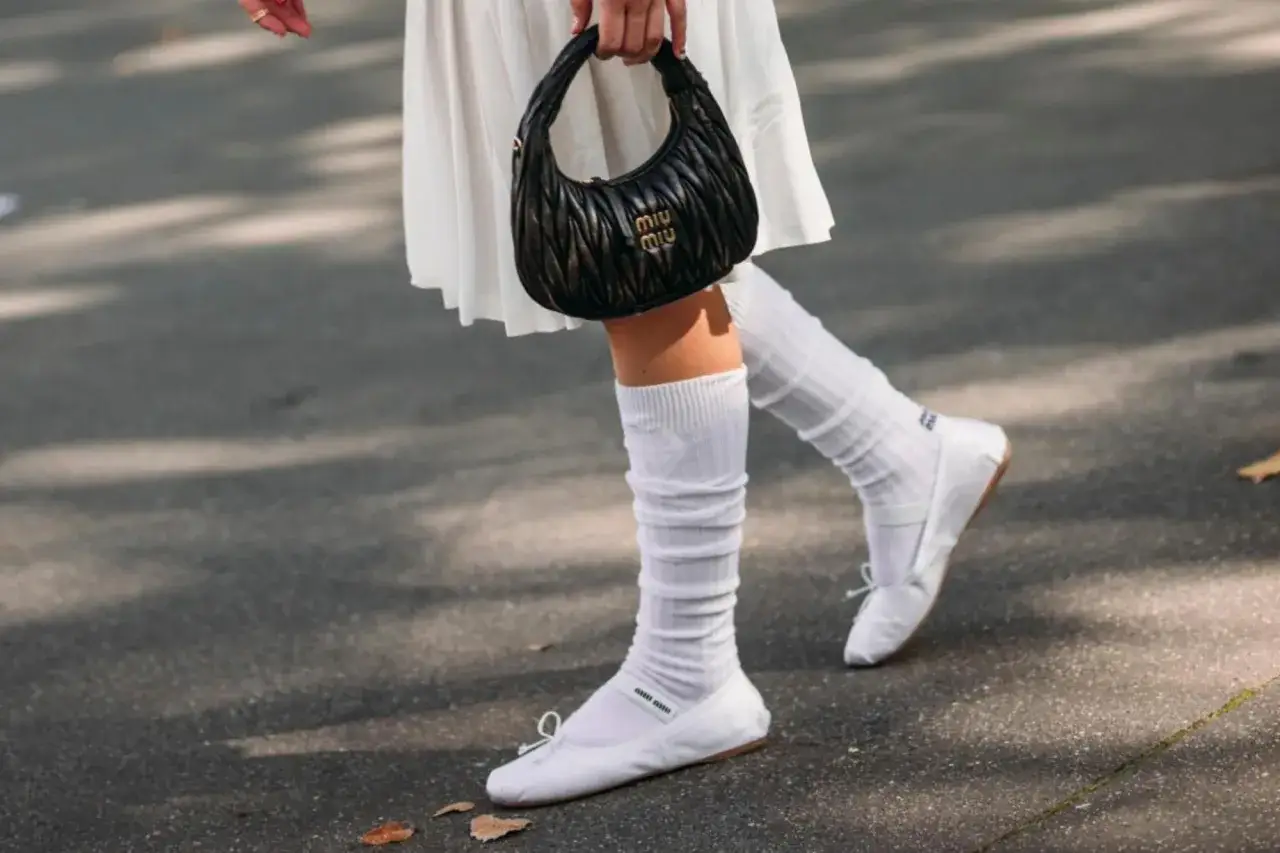 Piernas con bailarinas blancas y calcetines blancos arrugados, sosteniendo un bolso negro.