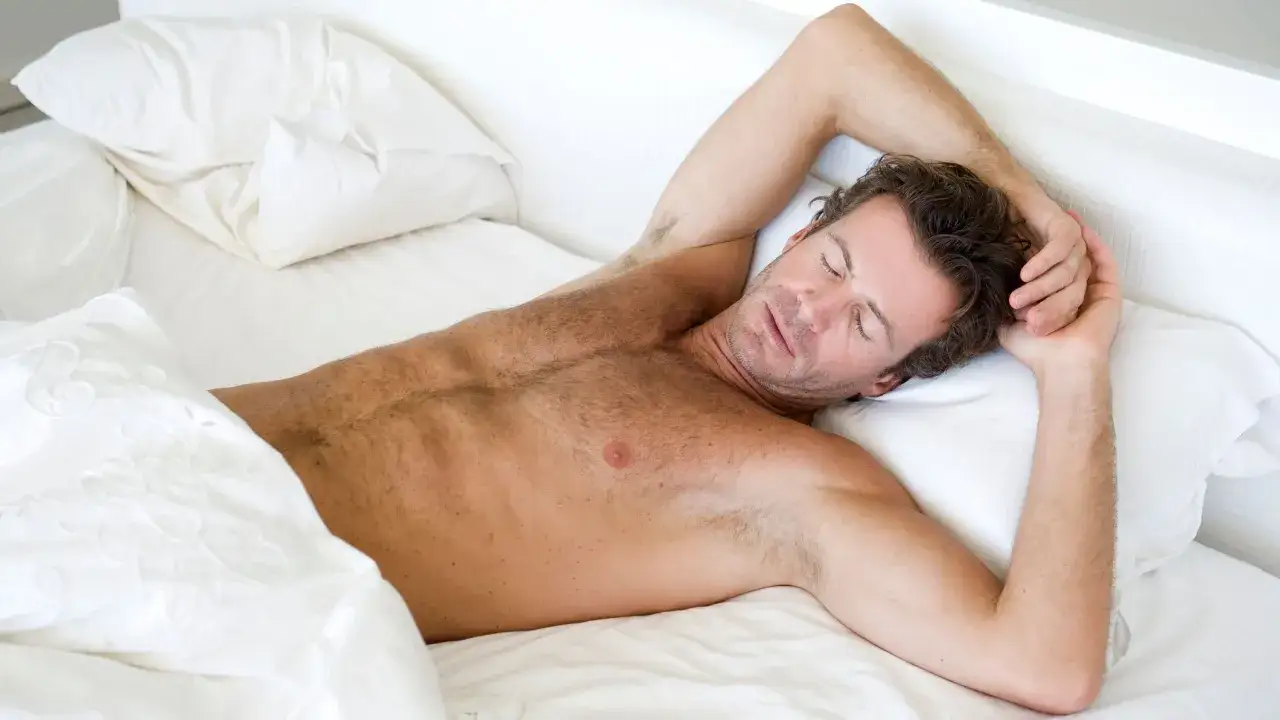 Hombre durmiendo pl&aacute;cidamente en una cama blanca, con los brazos cruzados sobre la cabeza. Se ve c&oacute;modo, quiz&aacute;s porque es bueno dormir sin ropa interior.