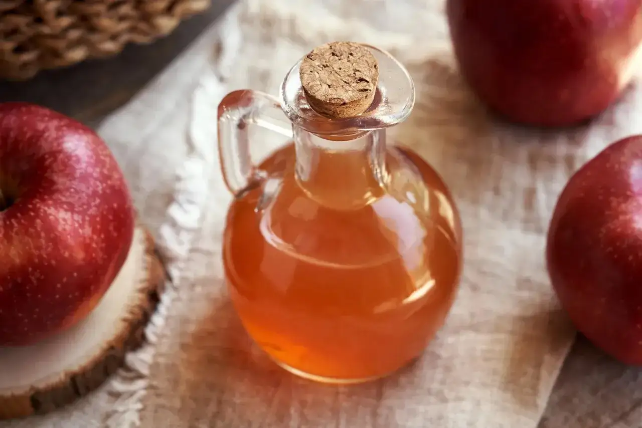Botella de vidrio con vinagre de sidra de manzana, rodeada de manzanas rojas frescas.